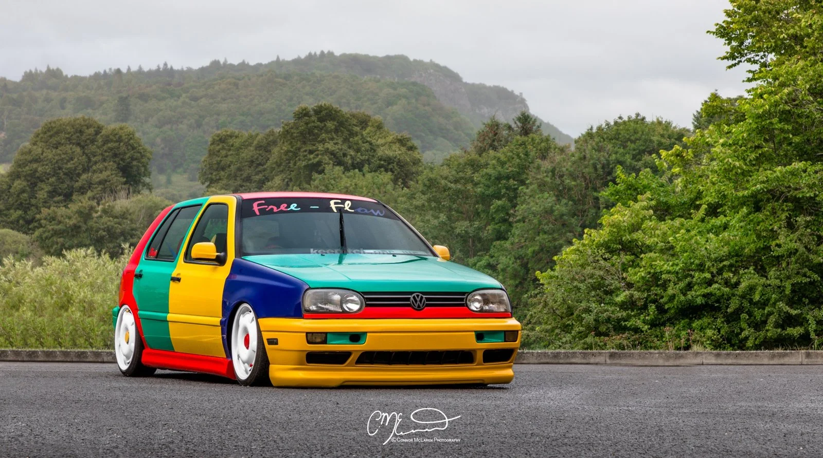 A colorful Volkswagen Golf Harlequin car with a rainbow paint job parked on a road near green trees and hills in the background.