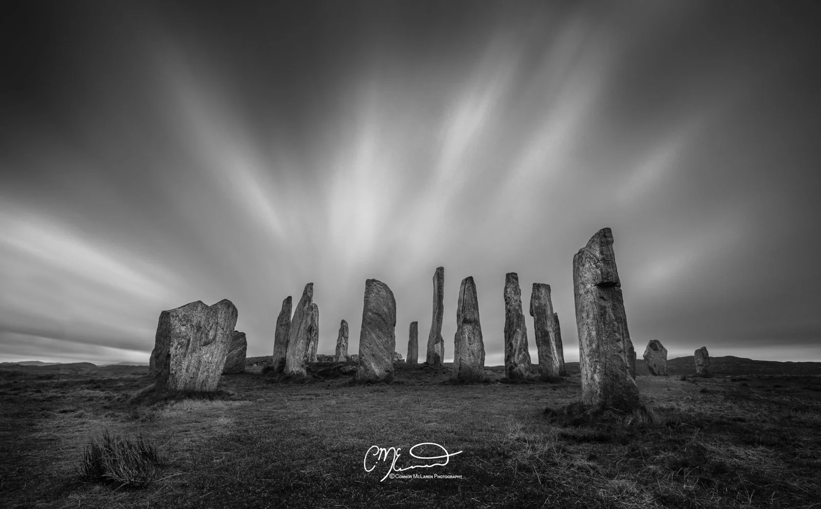 Scotland, Isle of Lewis Callanish Stones. Black and white photo of standing stones arranged in a circular pattern in a grassy landscape under dramatic sky with streaks of clouds.