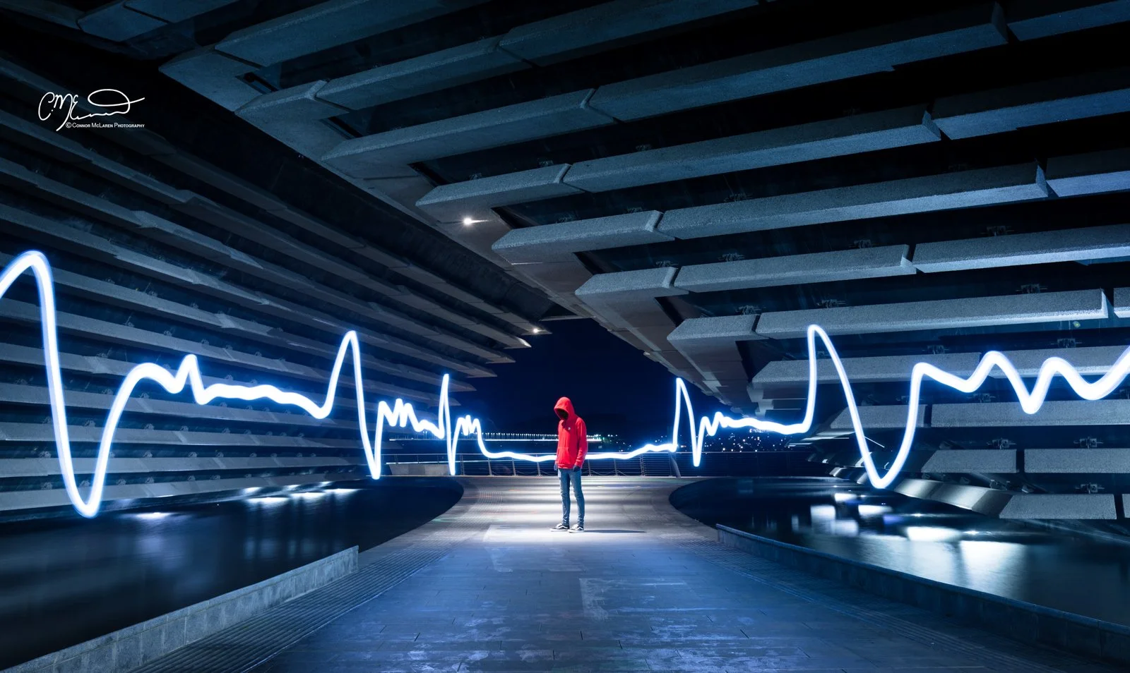 Dundee V&A, North East Scotland. A person in a red hoodie standing on an urban rooftop at night, with modern architecture and illuminated neon line resembling a heartbeat or waveform behind them.