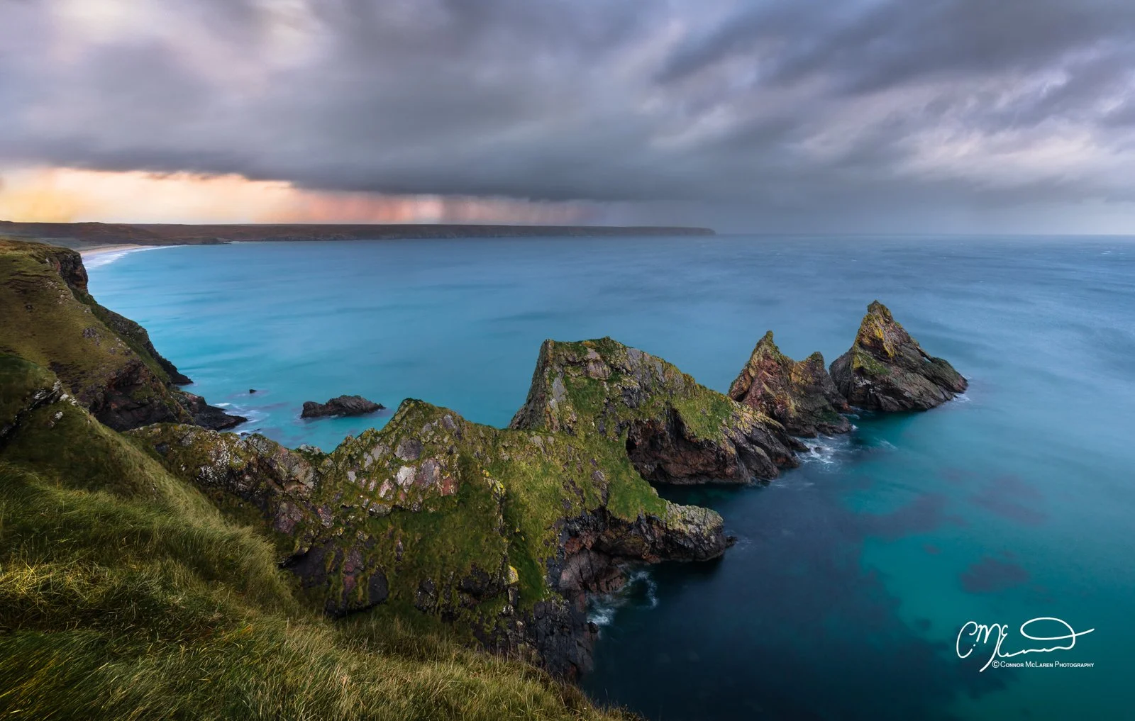 Scotland, Lewis Harris & Outer Hebrides, Tolsta. Cliffside view of rugged green mountains and rock formations overlooking a calm, blue ocean under a cloudy sky with a distant storm or rain in the horizon.
