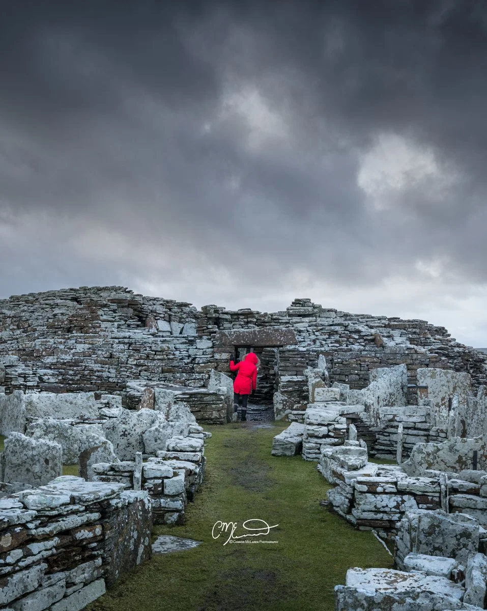Broch of Gurness, Orkney, Scotland. Person in a red coat walking through ancient stone ruins on a cloudy day.