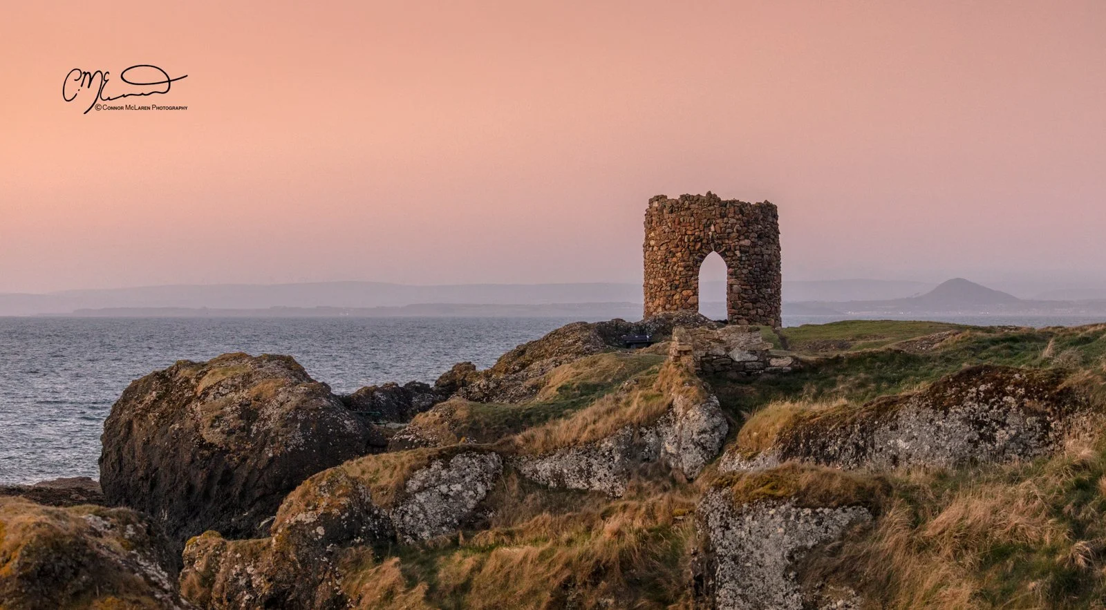 Scottish Lowlands, Scotland, east neuk of fife. Ancient stone tower on rocky grassy coast at sunset with calm water and distant mountains.