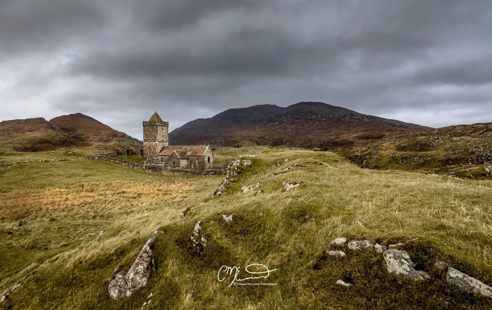 Scotland, Lewis Harris & Outer Hebrides. A stone church with a tower sits on a grassy hill with rocks, surrounded by rolling hills and mountains under a cloudy sky.