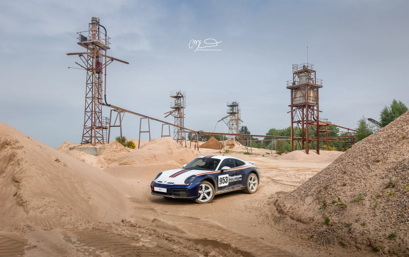 A white and blue Porsche 911 Dakar Roughroads race car with the number 953 on its side is parked on a sandy dirt track with sand dunes. In the background, there are rusted industrial towers and pipes under a cloudy sky.