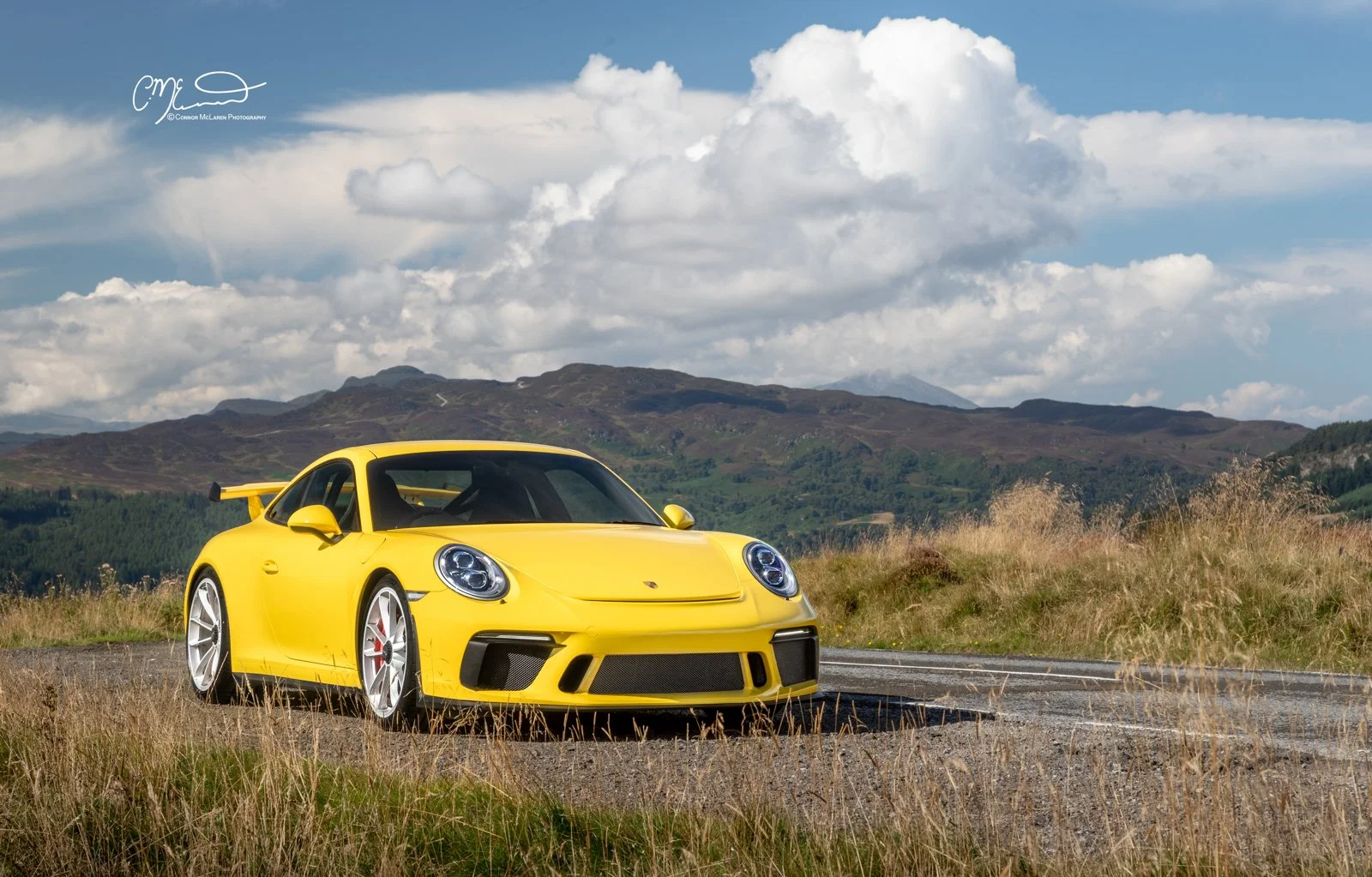 A yellow Porsche 911 GT3, 991 sports car on a rural road with mountains and a partly cloudy sky in the background.