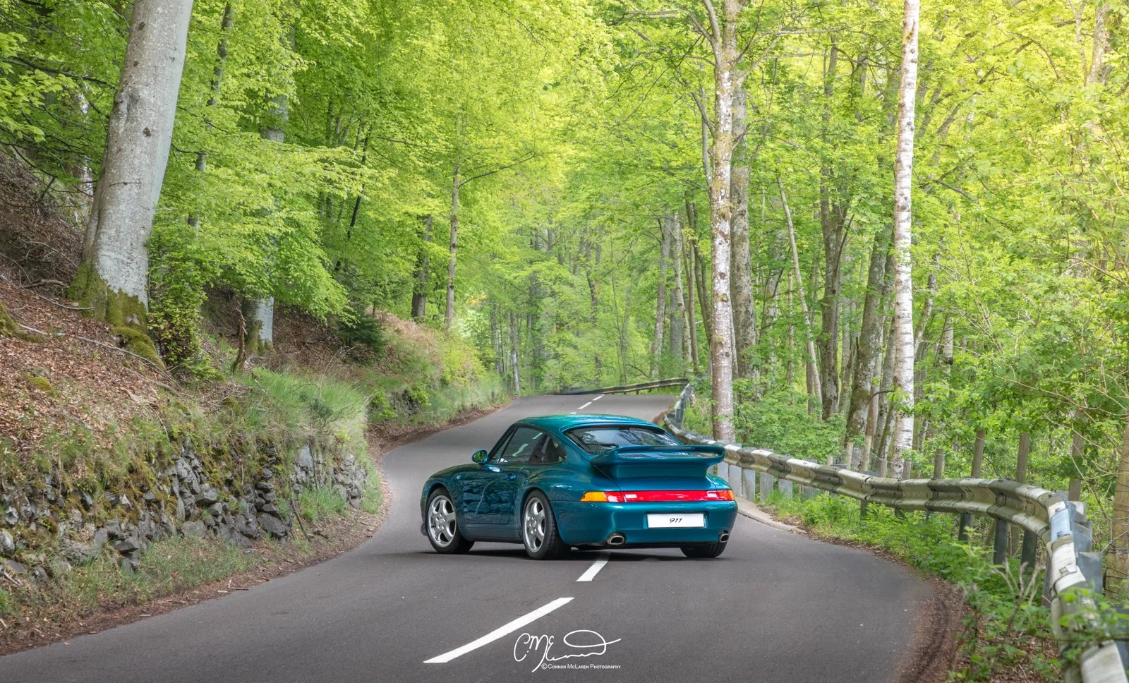 A blue Porsche 911 parked on a winding forest road surrounded by green trees and foliage.