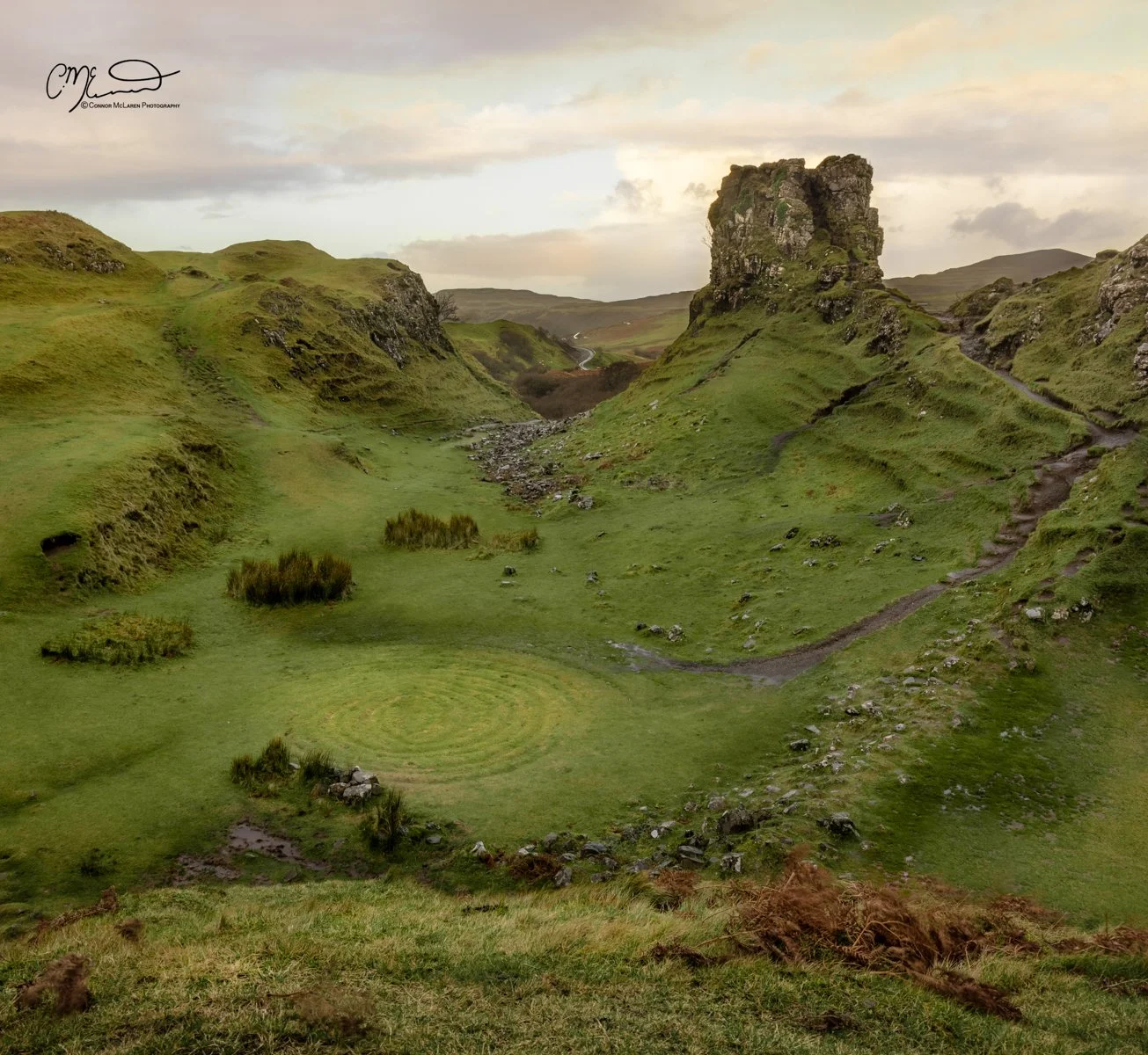 Scotland, Isle of Skye, fairy glen. A scenic landscape of green rolling hills with a distinct rock formation on the right, a winding path, and cloudy sky.