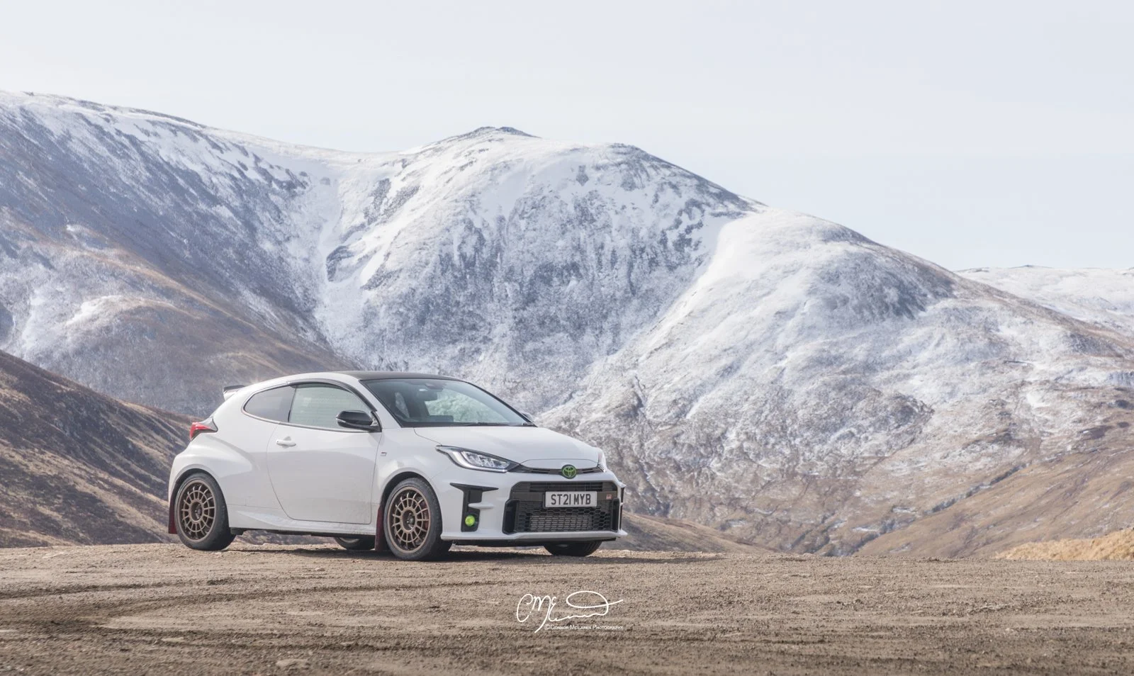 White Toyota Yaris GR hatchback car parked on a dirt road with snowy mountains in the background.
