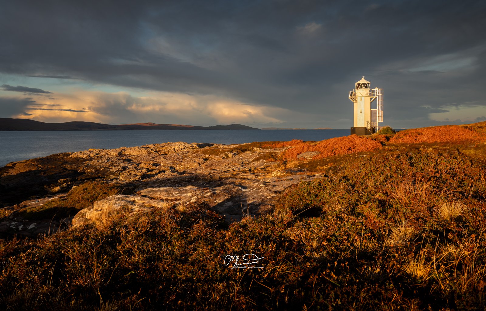 Scotland, north coast 500, NC500, Rhue Lighthouse. Lighthouse on rocky coastline during sunset with dramatic dark clouds in the sky and distant land across the water.