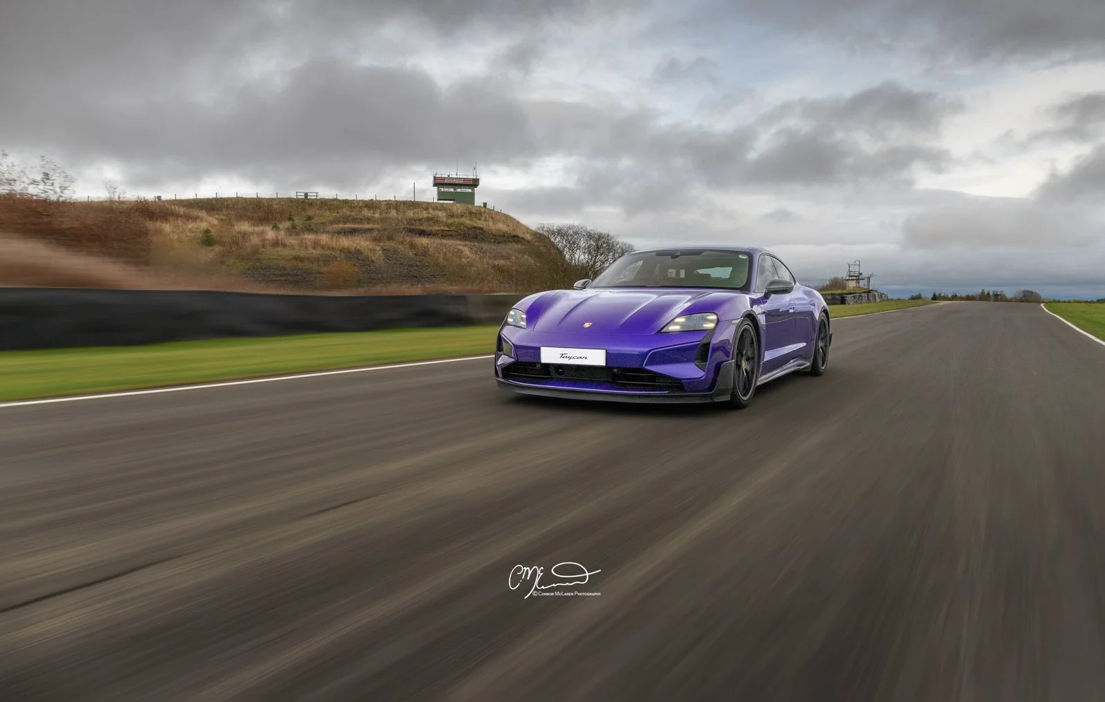 Porsche Taycan Turbo GT in purple racing at Knockhill. A purple sports car racing on a track under cloudy skies, with grassy hills and a watchtower in the background.