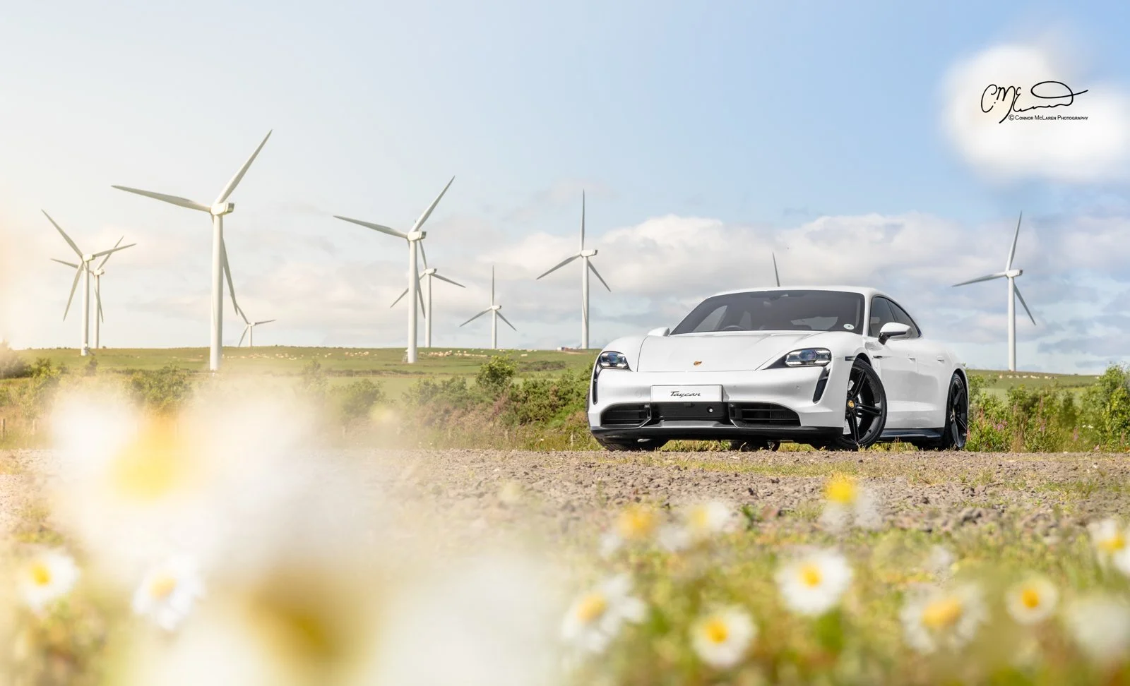 A white Porsche Taycan Turbo S parked on a dirt road with wind turbines in the background under partly cloudy sky.