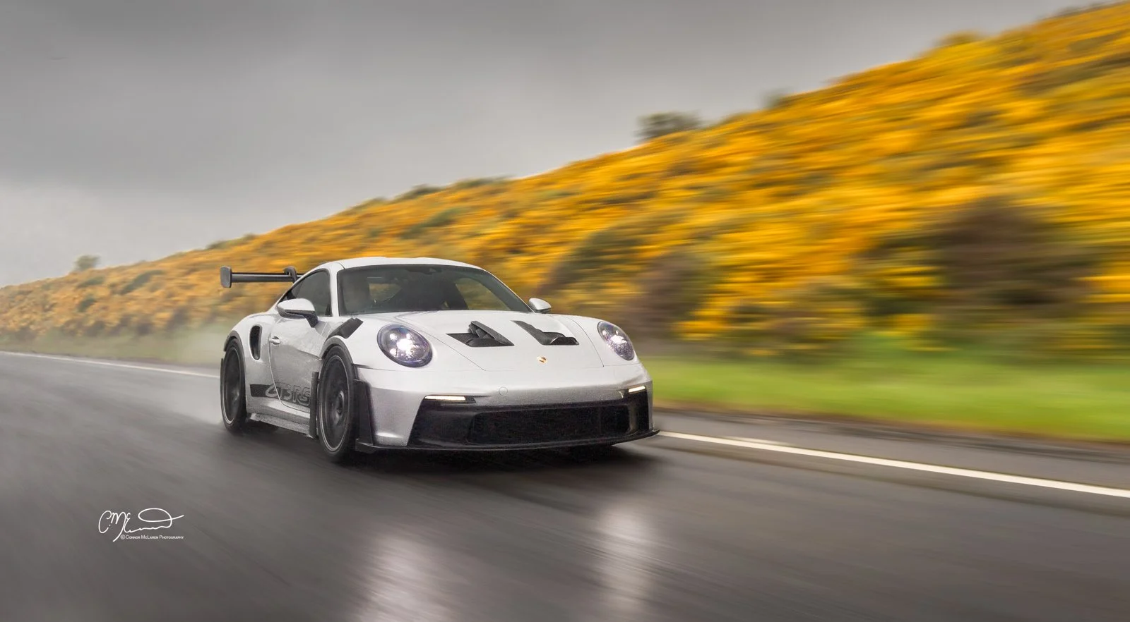 A silver Porsche 911 GT3 RS  racing car on a road with a blurred background of yellow flowers and gray sky.