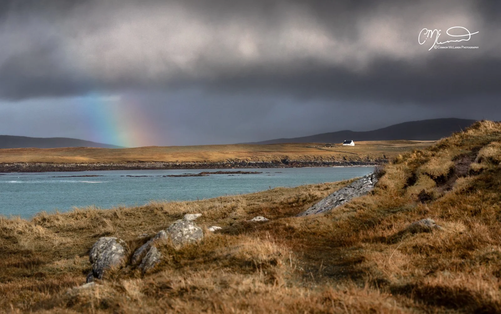 Scotland, Lewis Harris & Outer Hebrides, north Uist. A landscape with a large body of water, grassy hills, a solitary white house, a rainbow in the cloudy sky, and distant mountains.