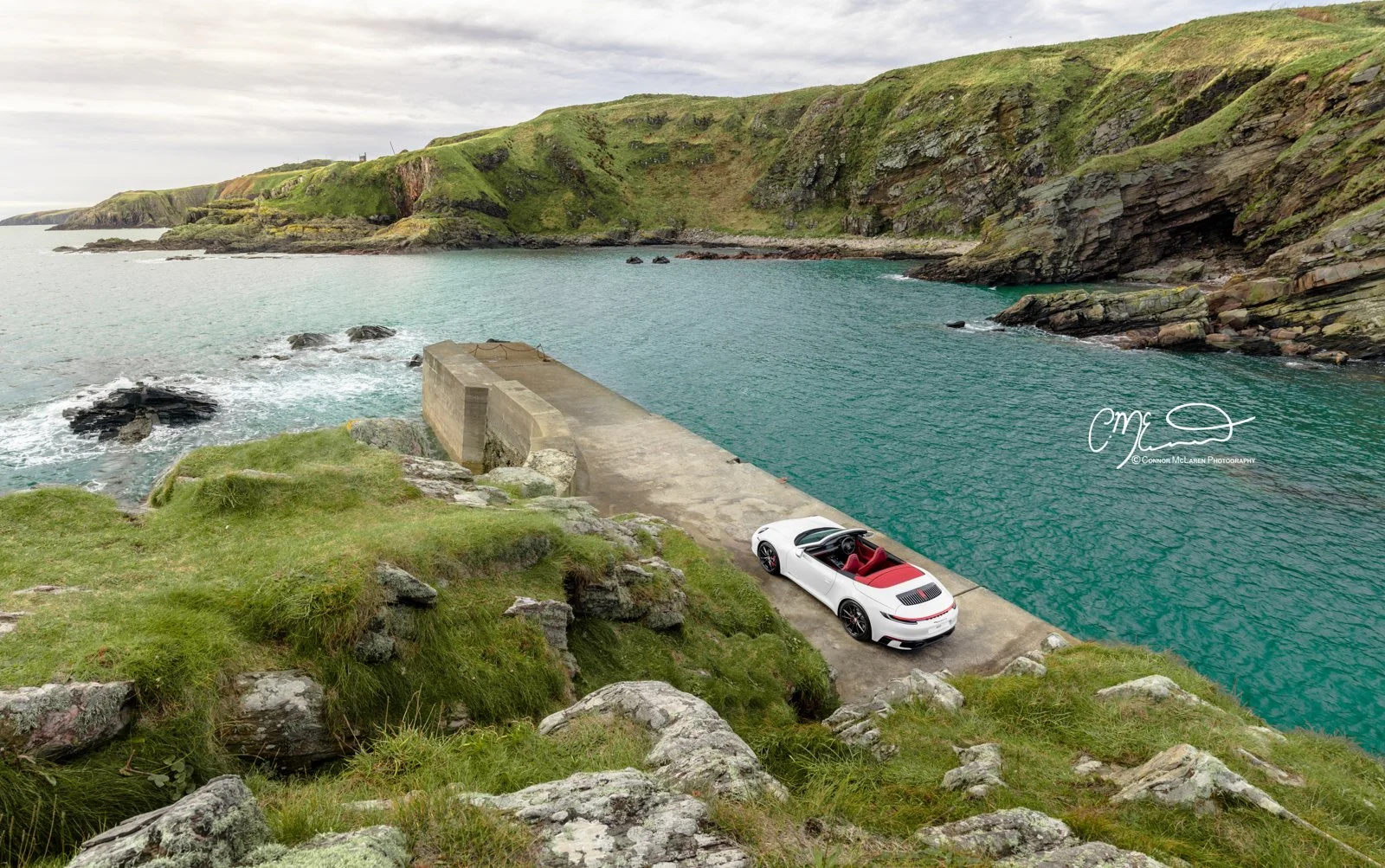 A white Porsche 911, 992 convertible car with a red interior parked on a concrete dock by the ocean, with green grassy hills and cliffs in the background.