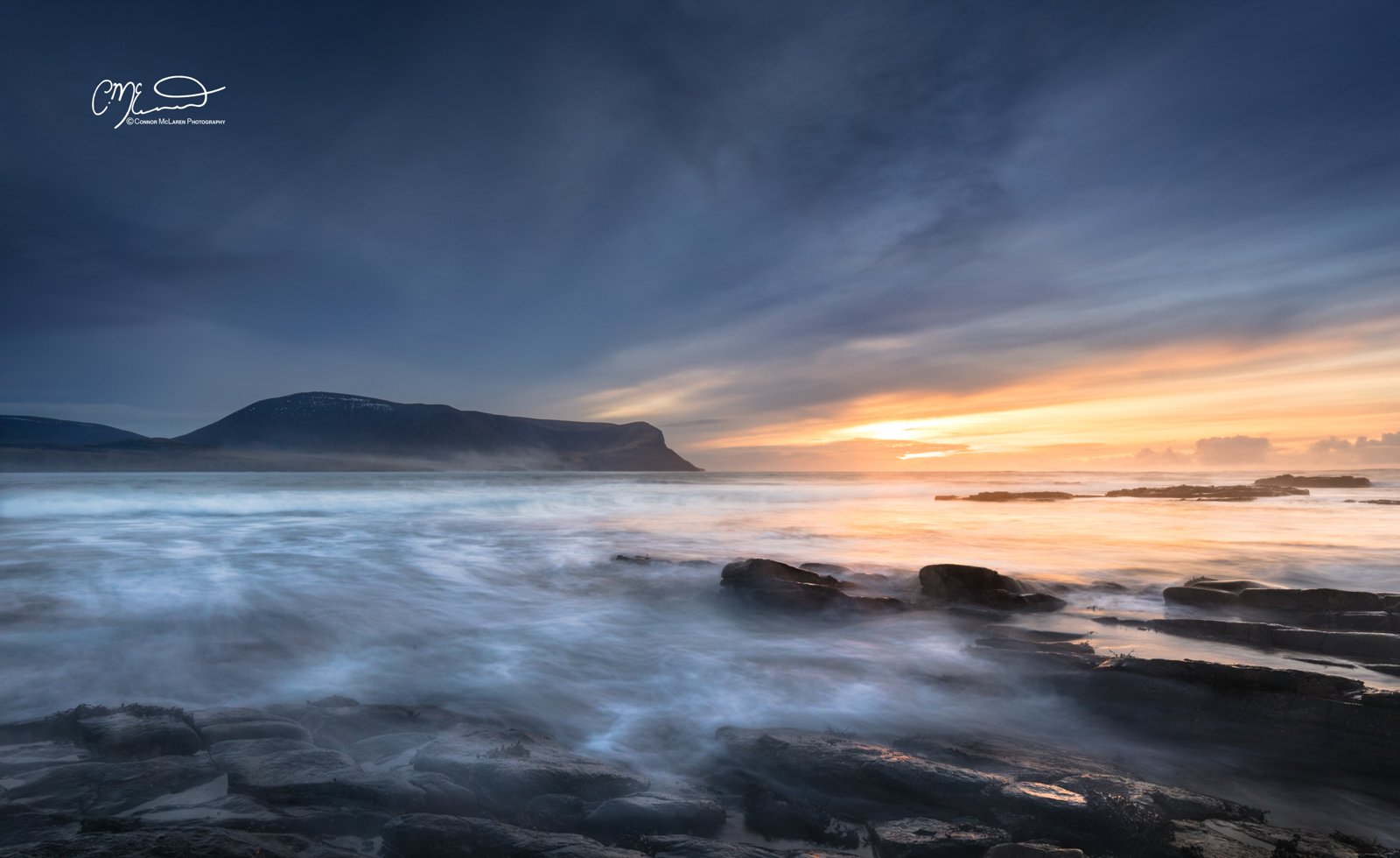 Orkney, Scotland. A scenic coastal landscape during sunset with a cloudy sky, rocky shoreline, and distant mountains.