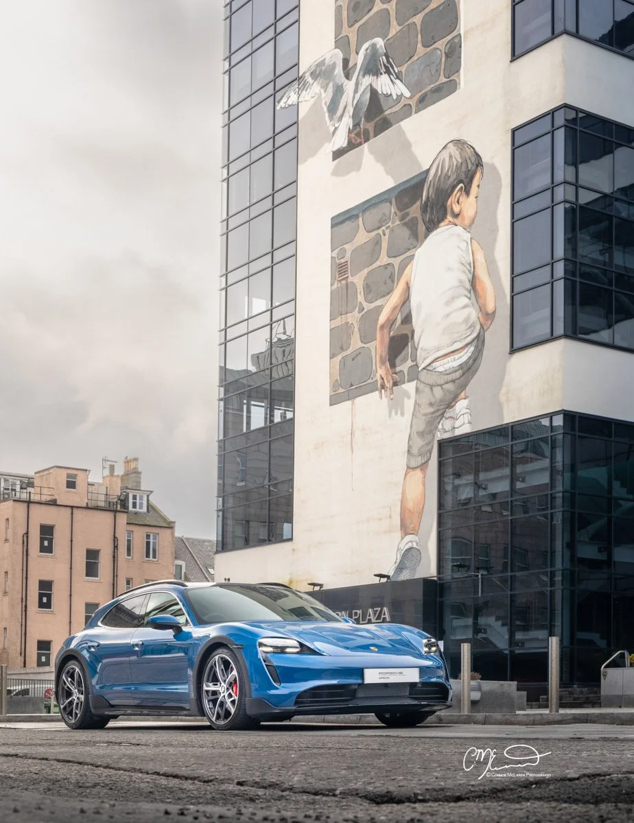 A blue Porsche Taycan Cross Turismo sports car parked in front of a modern building with a large mural of a boy climbing a wall and two pigeons flying above.