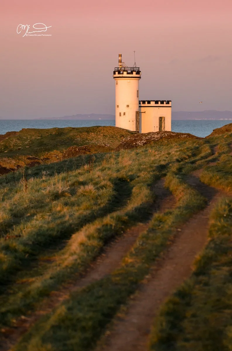 Scottish Lowlands, Scotland, east neuk of fife. A lighthouse situated on a grassy hill near the ocean during sunset or sunrise, with a dirt path leading towards it.