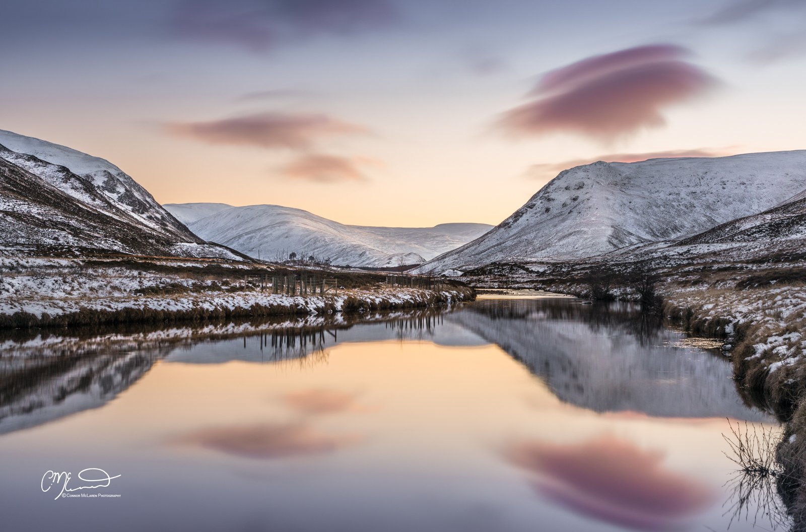 Scottish Central Highlands, Scotland, Perthshire Glen Shee. Snow-covered mountains surrounding a calm river that reflects the sky with pink, purple, and orange clouds during a sunset or sunrise.