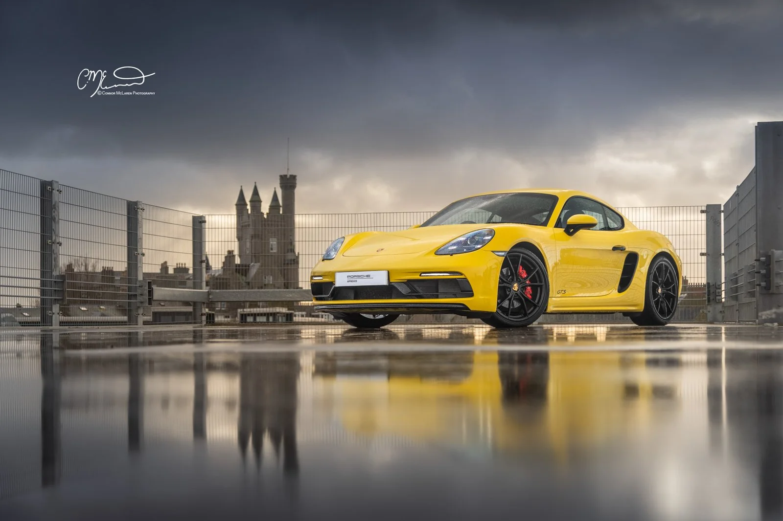 Yellow Porsche 718 Cayman GTS sports car parked on rooftop parking lot with cityscape and cloudy sky in the background