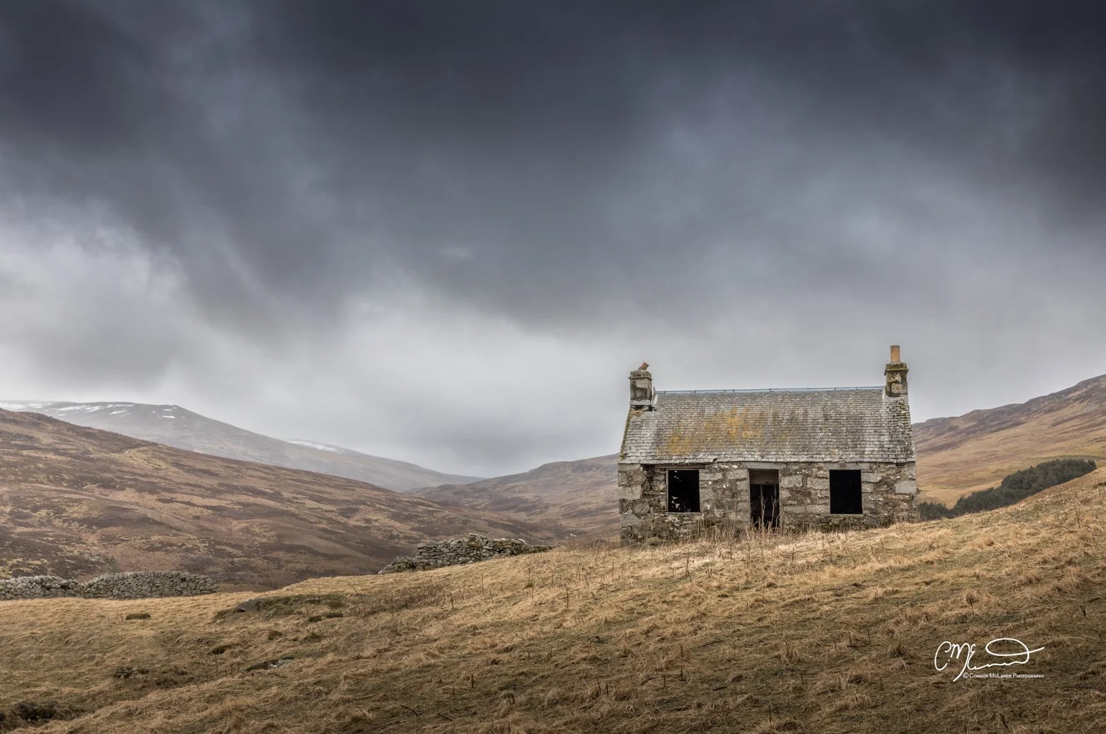 Scottish Central Highlands, Scotland, Perthshire bothy. A small, abandoned stone house with a pitched roof sits on a grassy hillside under dark, cloudy skies, with mountains in the distance.