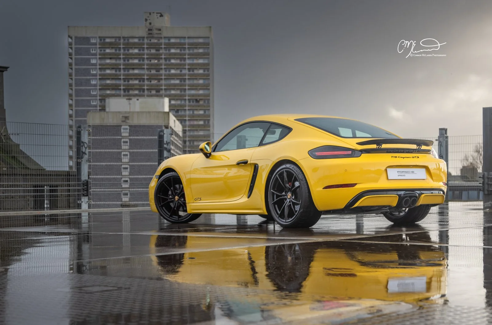 Yellow Porsche 718 Cayman GTS parked on top of a building with a cityscape background, reflections on wet ground.