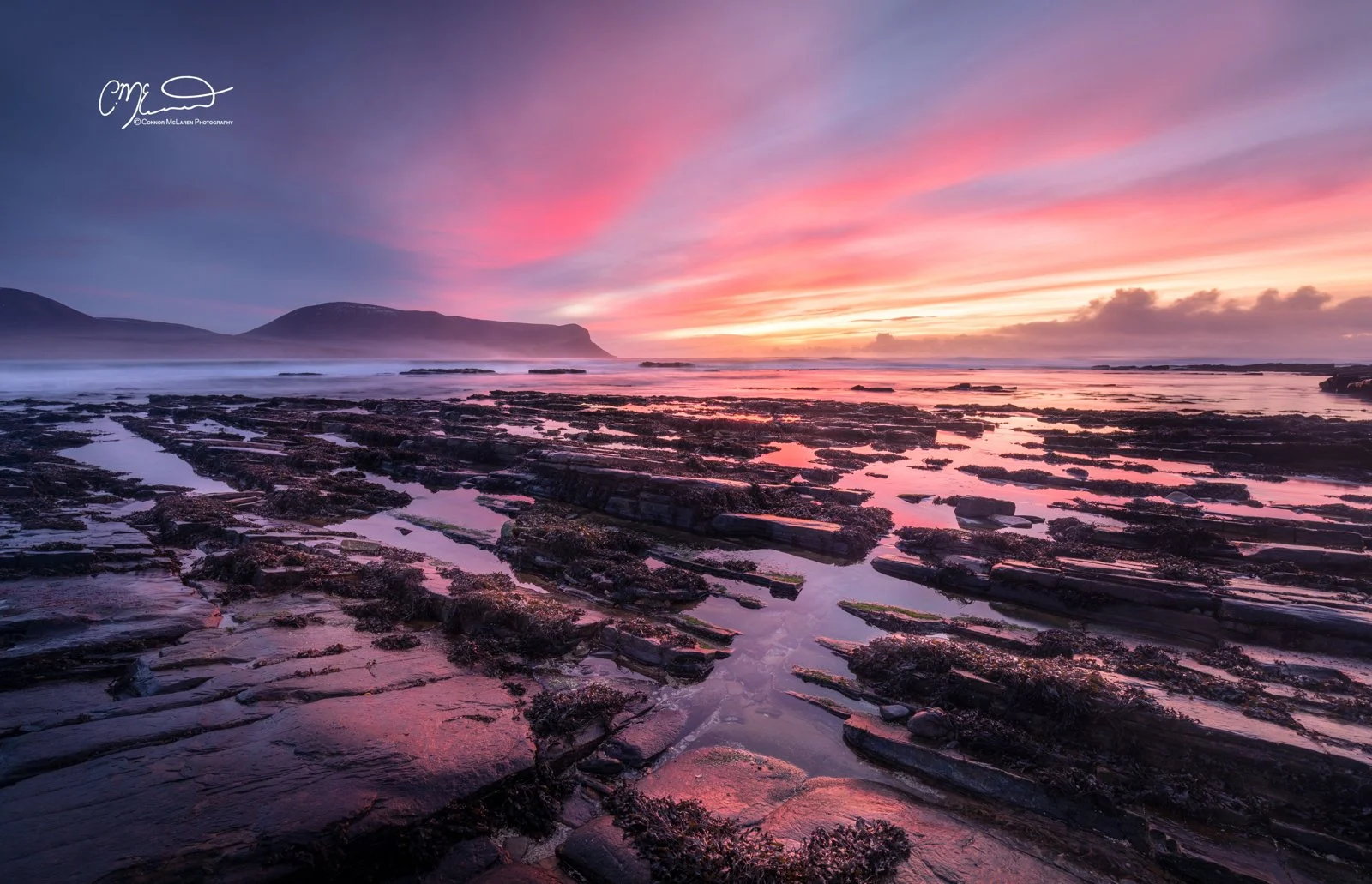 Orkney, Scotland. A scenic shoreline at sunset with pink and purple clouds, rocky tide pools, and distant mountains.