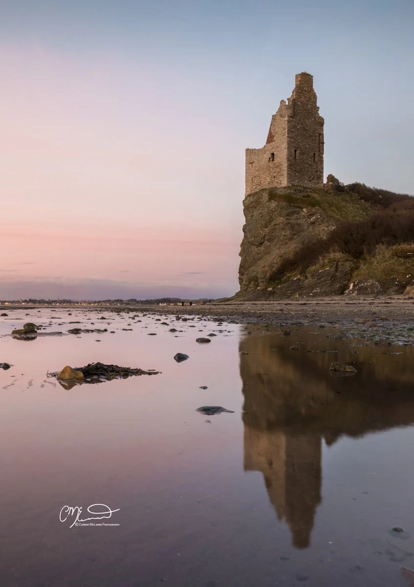 Scottish Lowlands, Scotland, greenan castle. A historic stone tower on a rocky cliff, reflected in a calm, shallow body of water at sunset.