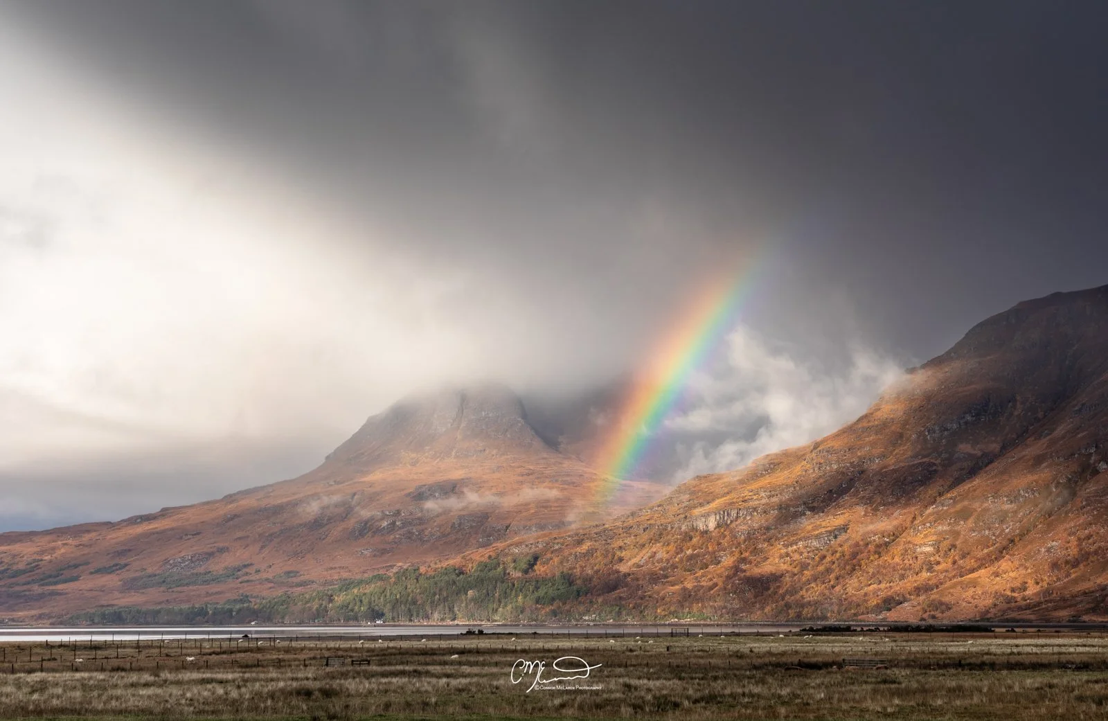 Scotland, north coast 500, NC500. A rainbow arches over a mountainous landscape with cloudy skies and autumn-colored slopes.