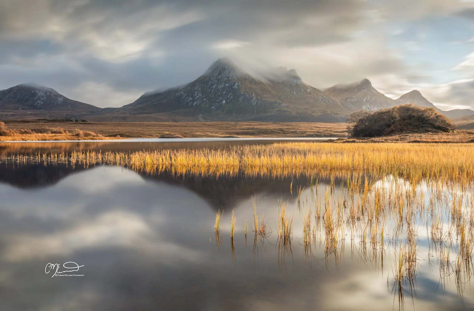 Scotland, north coast 500, NC500, Lochan hakel, ben loyal. Scenic view of a mountain range with mist-covered peaks reflected in a calm lake, surrounded by grassy fields and trees in warm, autumn colors.