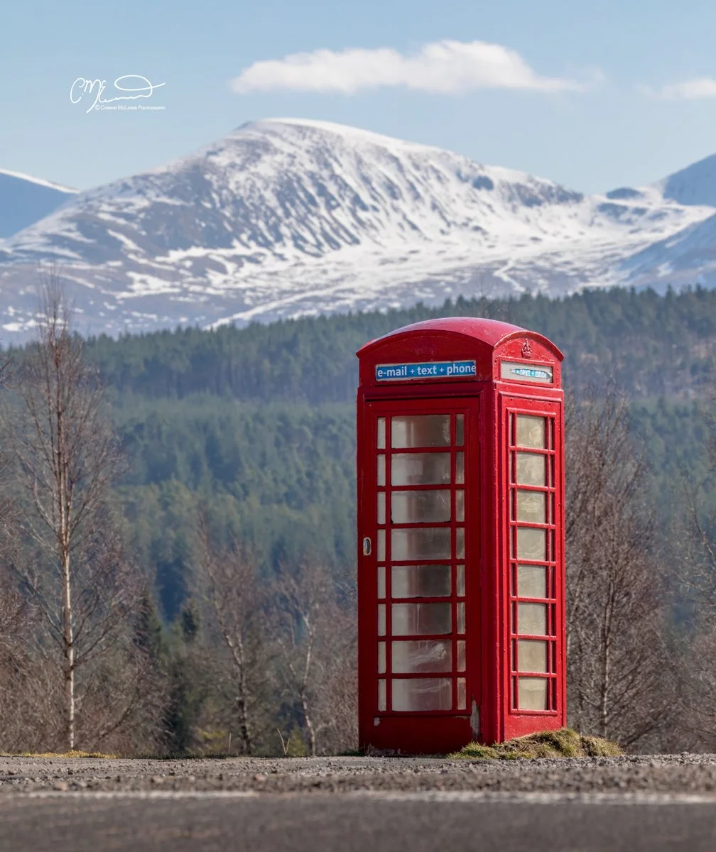 Scottish Central Highlands, Scotland, Perthshire. A red old-fashioned telephone booth standing on a road with snow-capped mountains in the background and some leafless trees around.