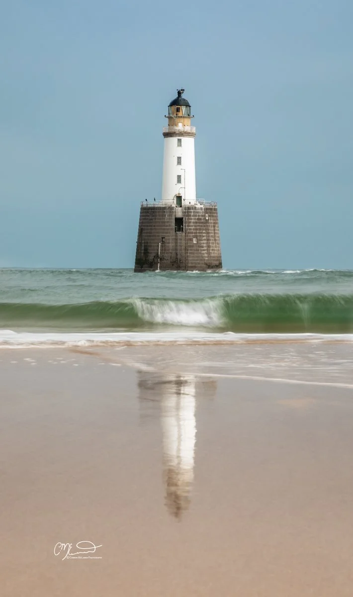rattray head lighthouse, rainbow, North East Scotland. A lighthouse on a small rocky island in the ocean, with waves lapping at the shore and a reflection of the lighthouse in the wet sand.