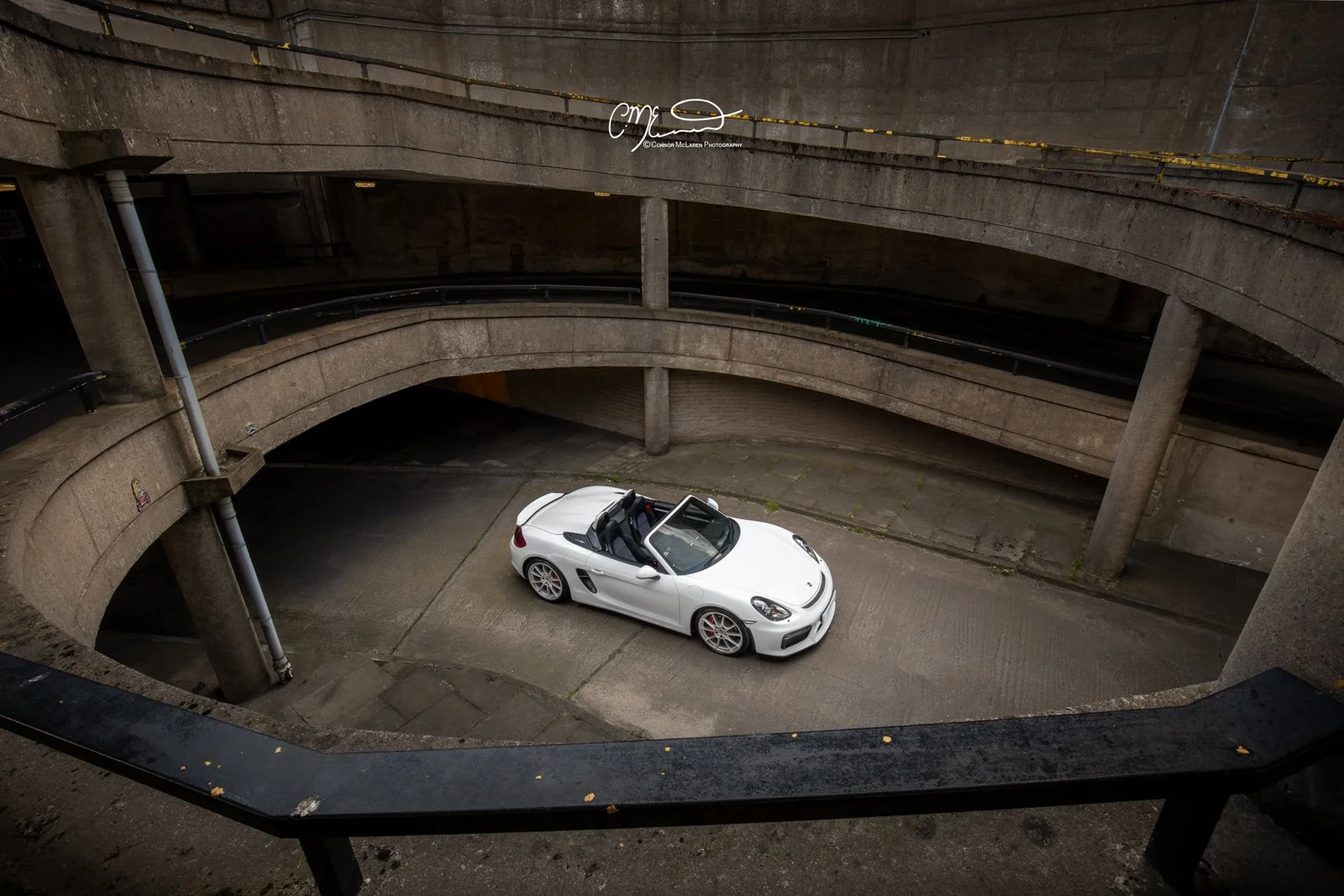 White Porsche Boxster Spyder convertible sports car parked in an underground parking garage with concrete walls and spiral ramps.