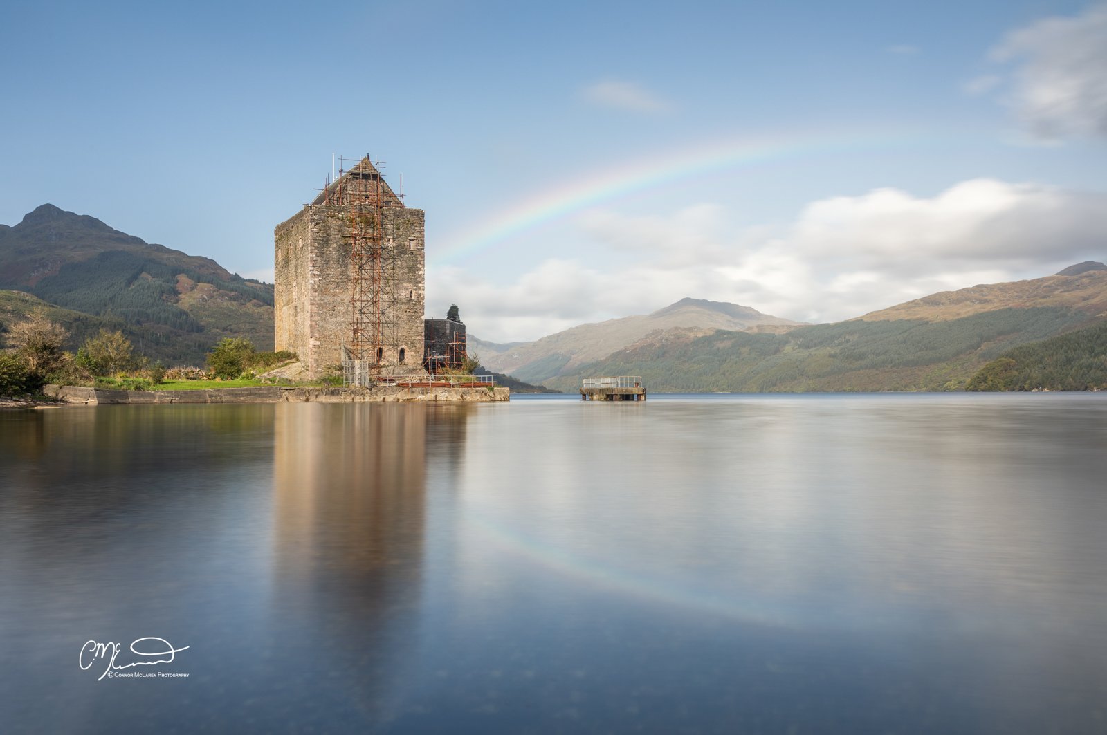 Castle Carrick, Rainbow, Western Highlands, Scotland. An ancient stone tower on the shoreline of a lake, with mountains in the background and a rainbow in the sky.