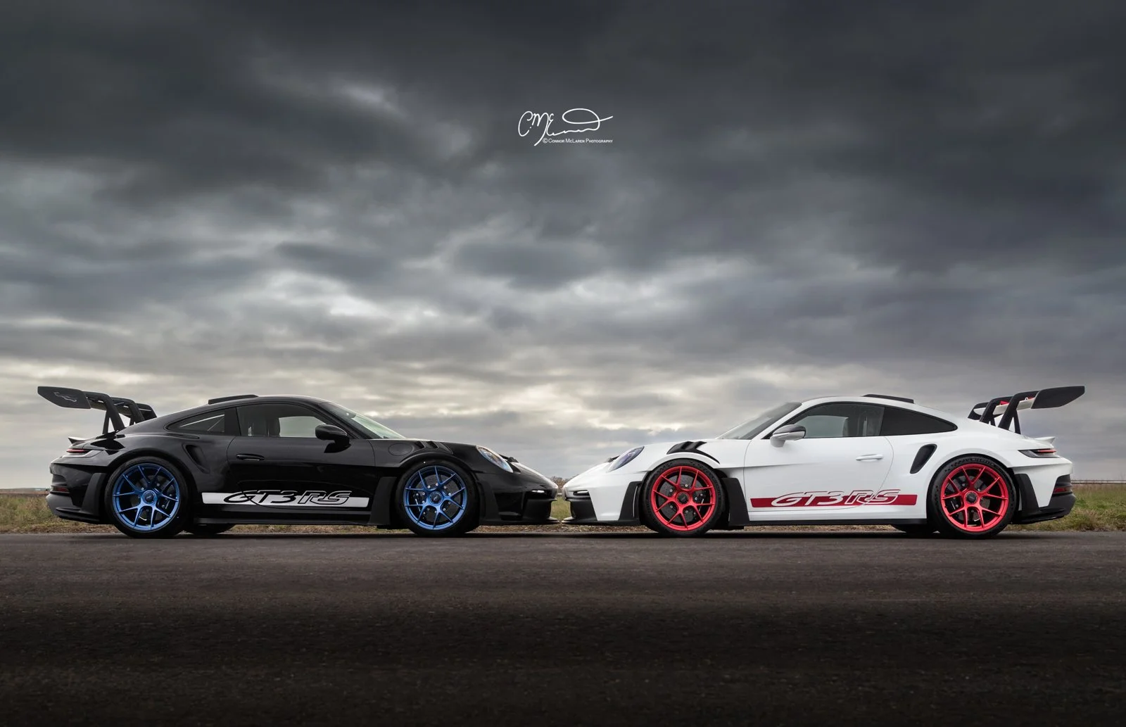 Two Porsche 911 GT3 RS cars parked on a road under a cloudy sky, one black with blue wheels and the other white with red wheels, both with prominent rear wings and rally stripes.