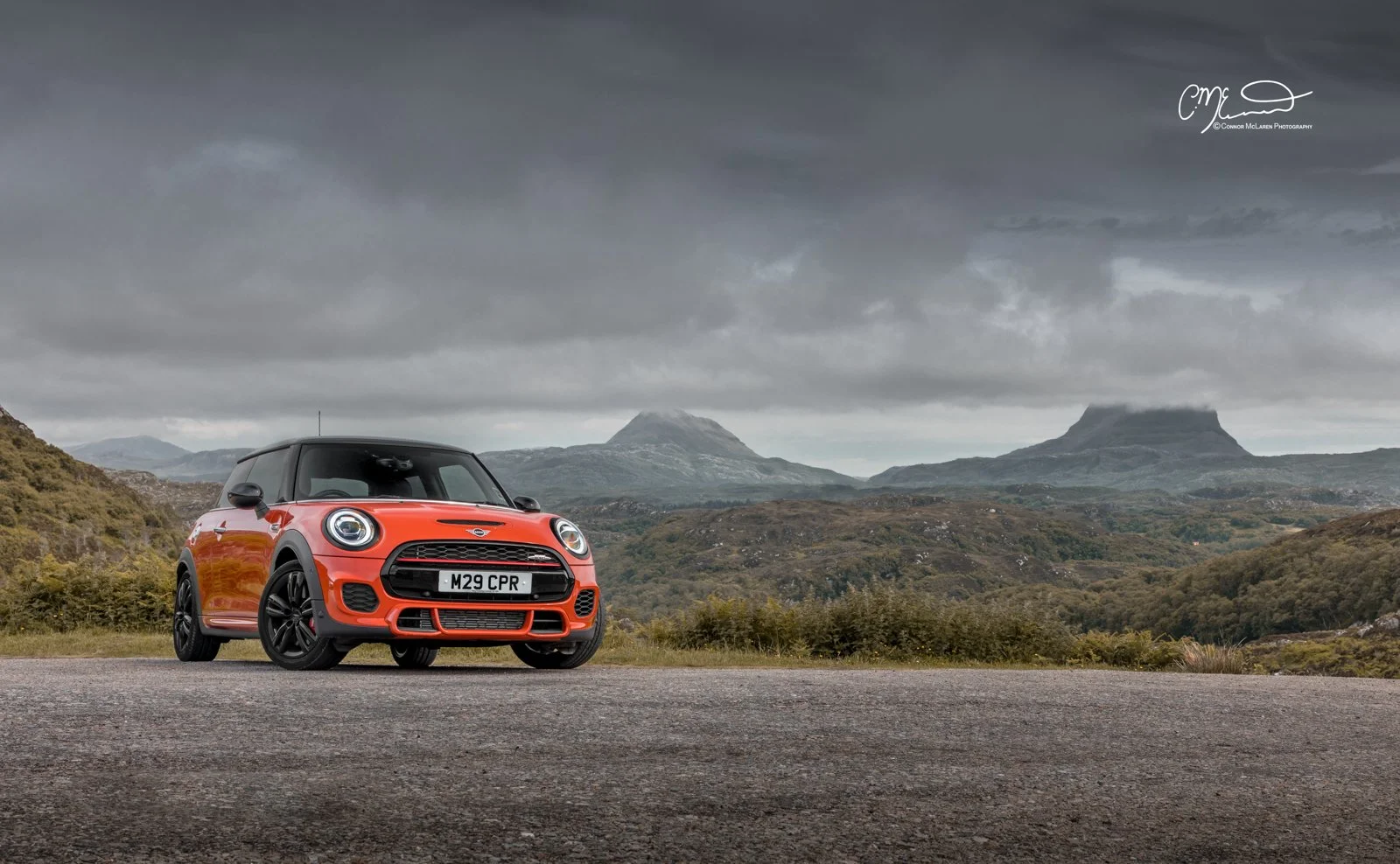 A red and black Mini Cooper parked on a gravel road in a mountainous landscape with dark cloudy sky.