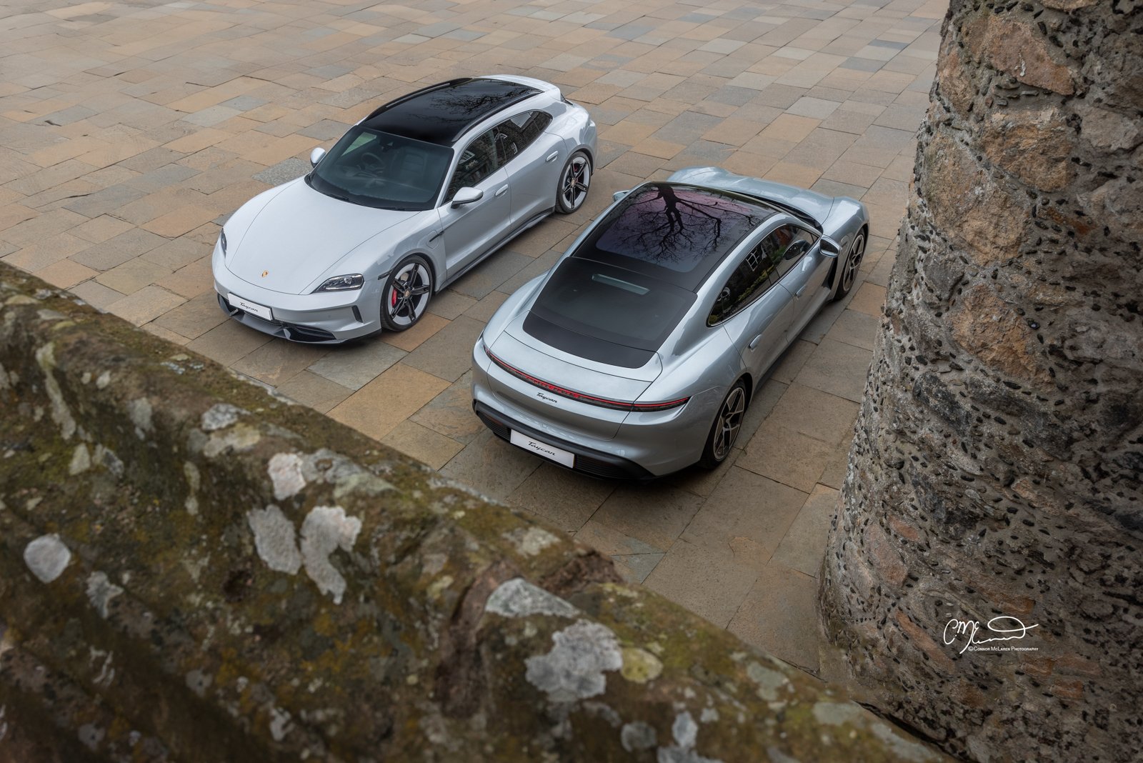 Two silver Porsche Taycan cars parked on a stone-paved surface, viewed from above between two stone walls.