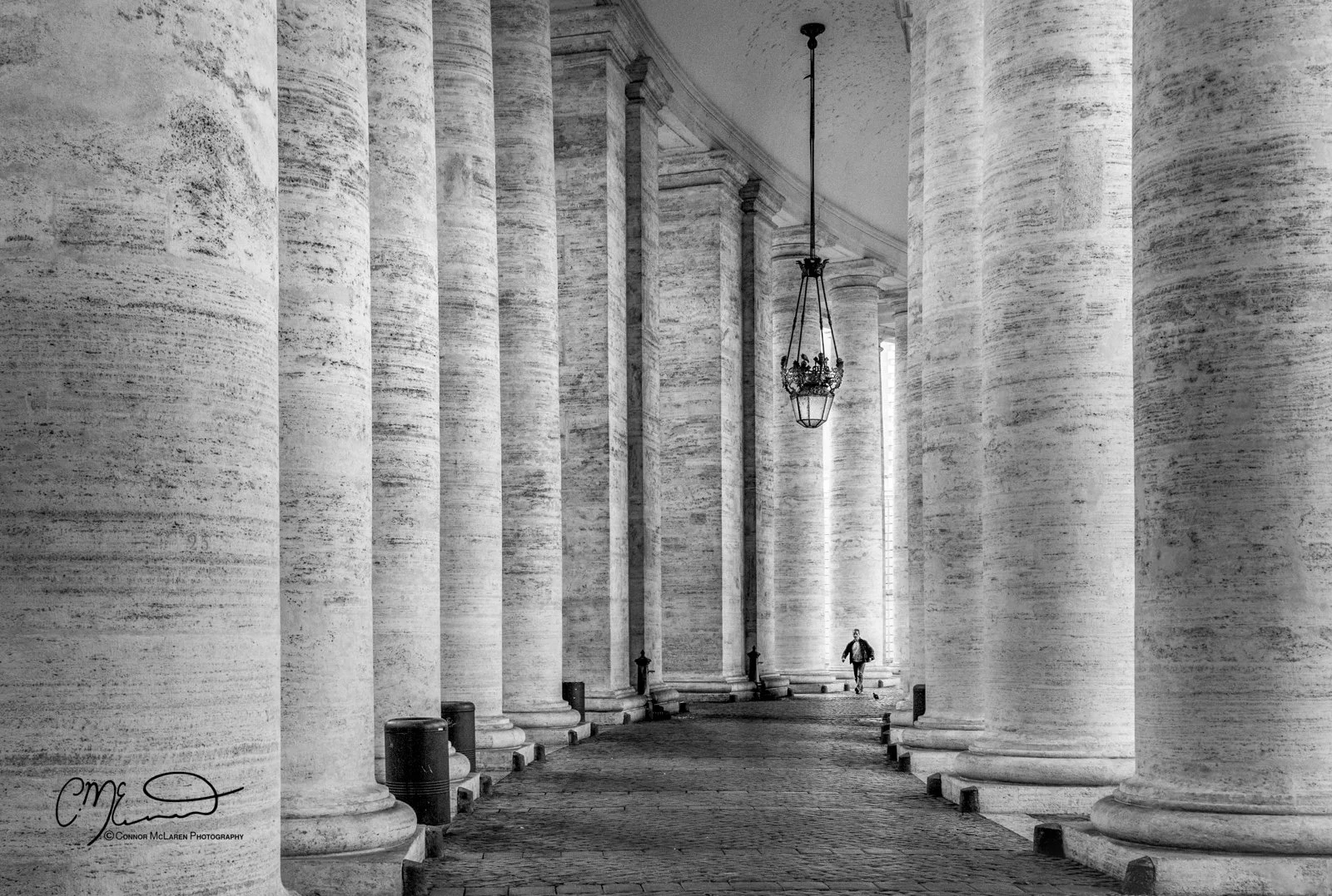 Black and white photograph of large stone columns forming a corridor, with a person walking in the distance. The columns have a textured appearance and are illuminated by natural light from the right side. Hanging from the ceiling is an ornate lantern-style light fixture.