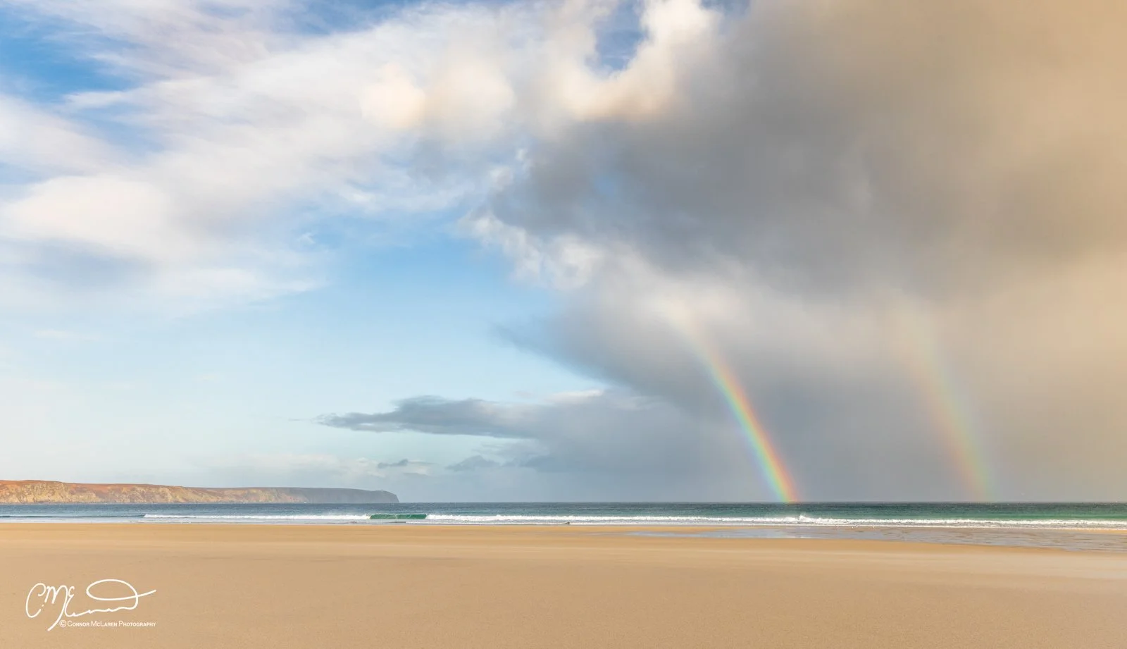Scotland, Lewis Harris & Outer Hebrides, Tolsta. A beach with a sandy shoreline, the ocean with small waves, a cloudy sky with rainbows, and cliffs in the distance.
