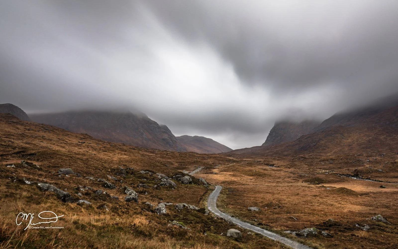 Scotland, Lewis Harris & Outer Hebrides. A rugged landscape of rolling hills and mountains under a cloudy, overcast sky with a winding dirt path in the foreground.