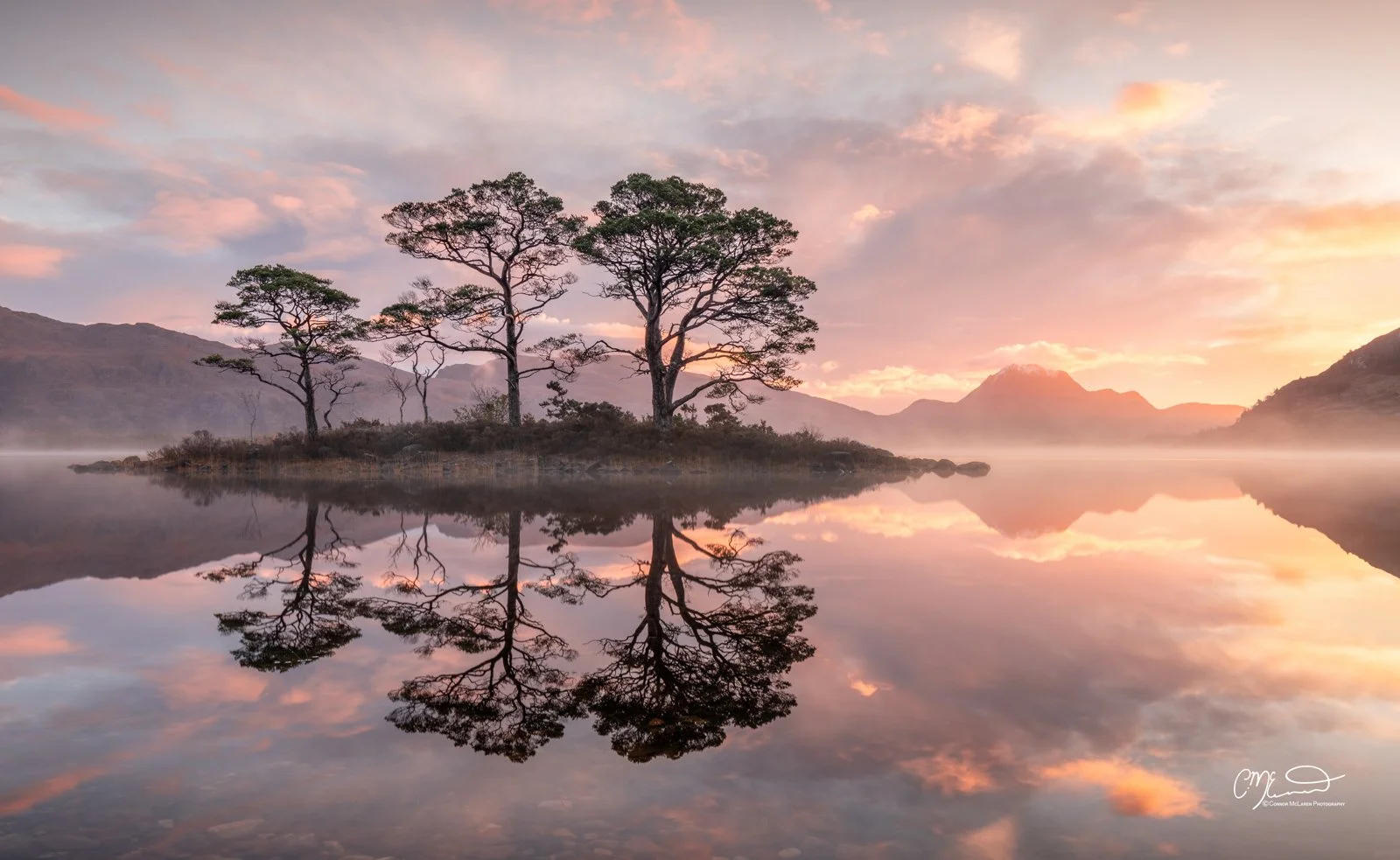 Scotland, north coast 500, NC500, Loch Maree. A serene landscape at sunrise with a small island of trees reflected in calm water, surrounded by mountains and a colorful sky with pink and orange clouds.