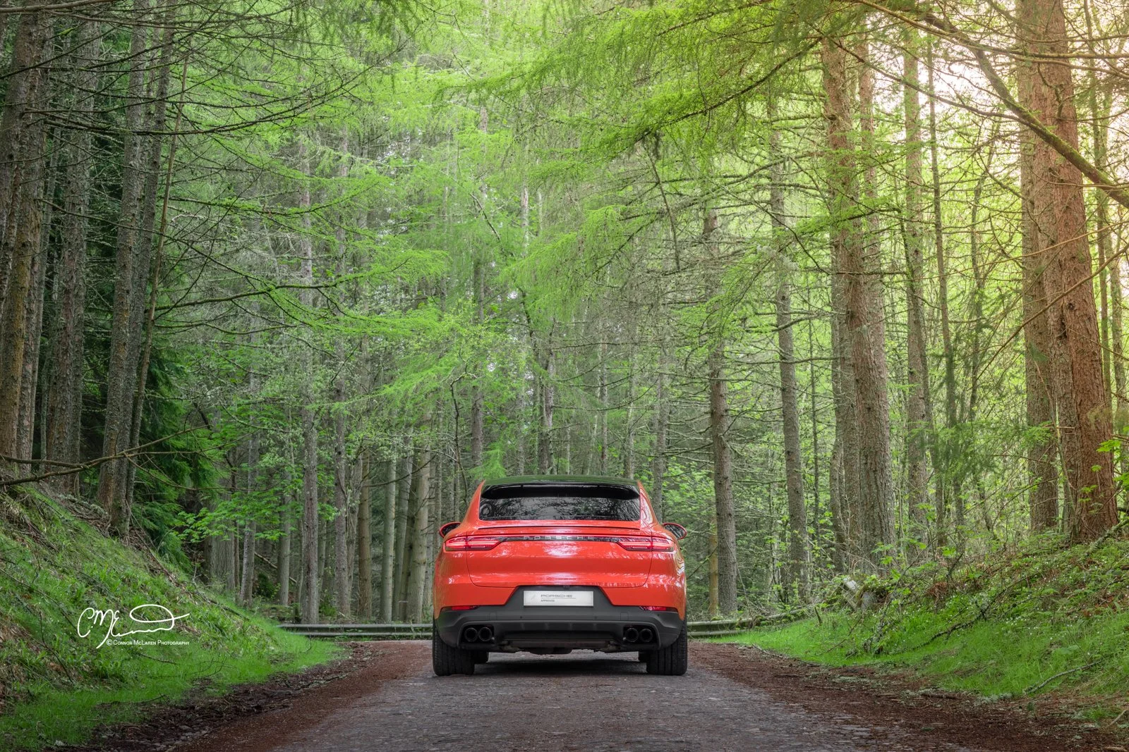 Lava Orange Porsche Cayenne Coupe. Red car driving on a dirt forest trail surrounded by green trees and foliage.