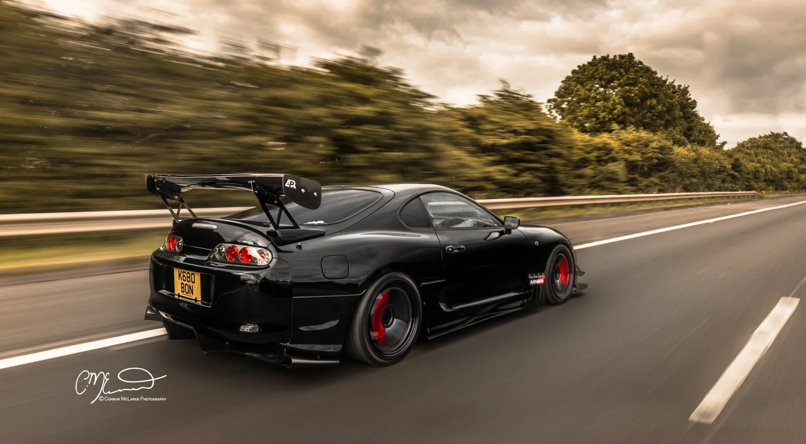 Custom Modified Toyota Supra 2JZ.A black sports car with red rims driving on a highway at sunset, with trees and cloudy sky in the background.
