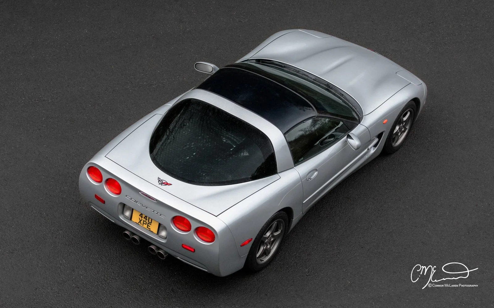 An overhead view of a silver Chevrolet Corvette sports car parked on a dark asphalt surface. The car features a black roof, round taillights, dual exhaust pipes, and a license plate reading 440 XPE. The Corvette logo is visible on the rear of the vehicle.