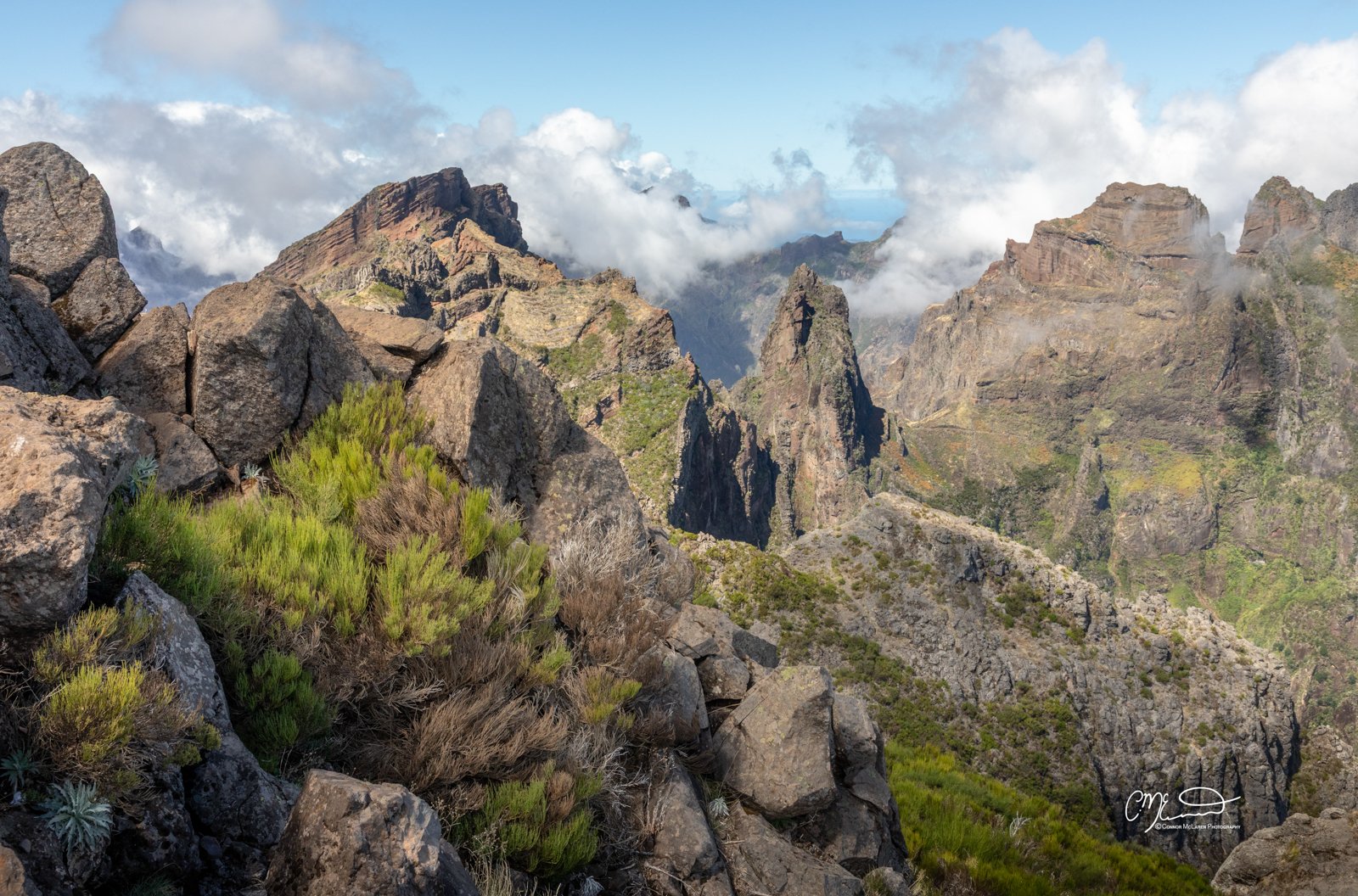 mountain top ridge, Madeira Portugal. Mountain landscape with large rocks and green vegetation in the foreground, rugged mountains in the background, and clouds in the sky.