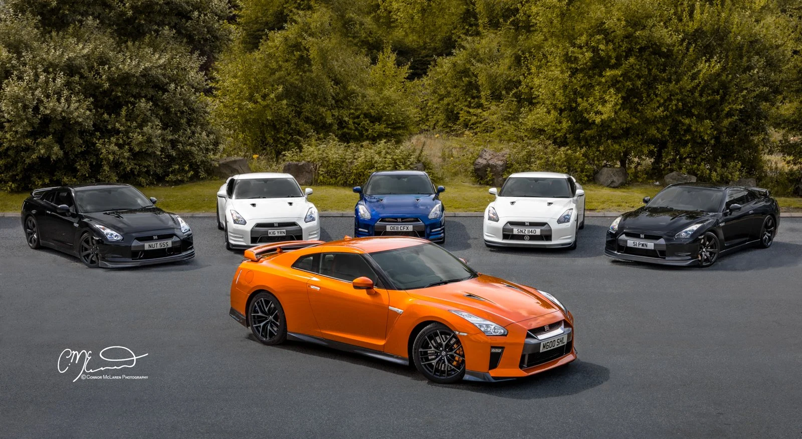 An orange Nissan GT-R sports car parked in front of six other cars in a line, with trees and bushes in the background.