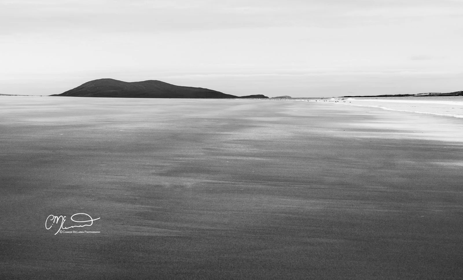 Scotland, Isle of Skye Bridge. Scotland, Isle of Harris Luskentyre Beach. A black and white landscape photo of a calm body of water with a distant hilly island or landmass on the horizon. The sky is mostly overcast with some clouds.