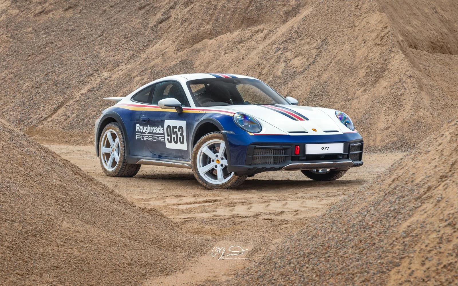 A Porsche 911 Dakar Roughroads painted in white, blue, and red racing stripes, with racing decals including 'Roughroads Porsche' and the number 953, parked on a sandy dirt track surrounded by mounds of dirt.
