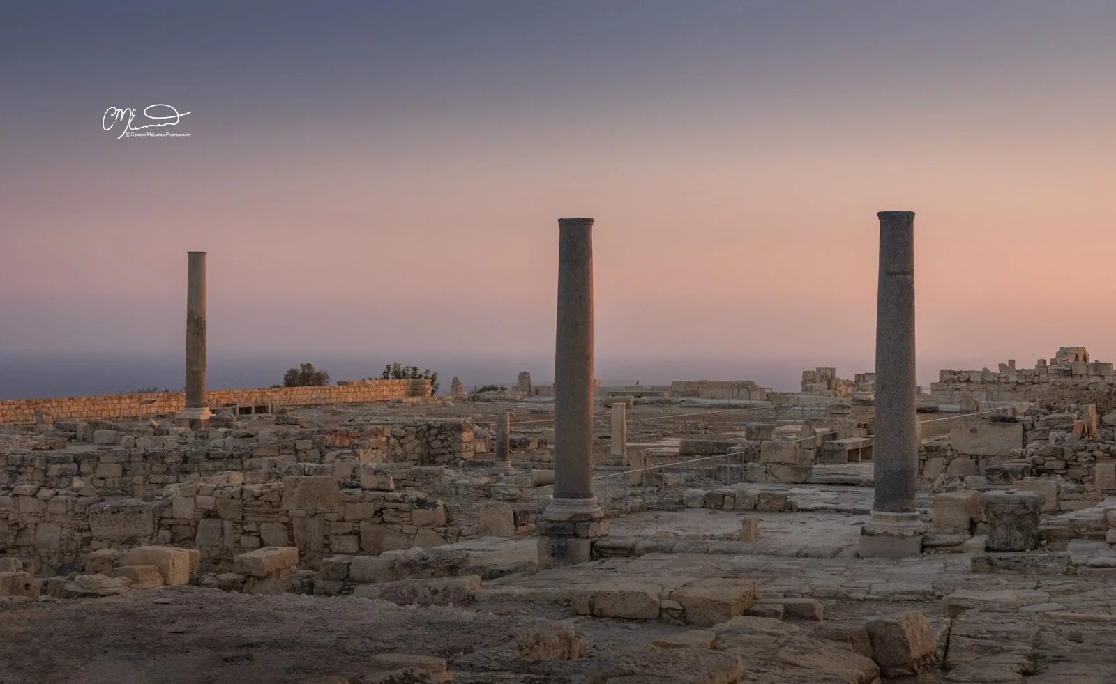 kourion, Cyprus. Ancient ruins with three stone columns and scattered stones, under a pastel gradient sky at sunset or sunrise.
