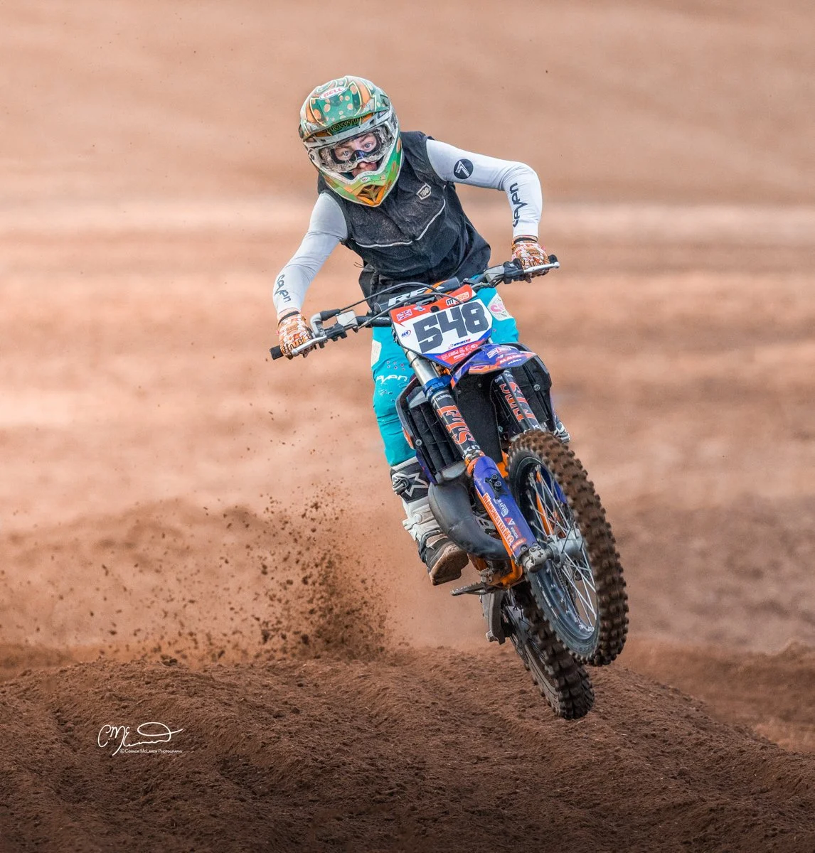 A motocross rider in full gear riding a dirt bike on a dirt track, airborne over a bump during a race.