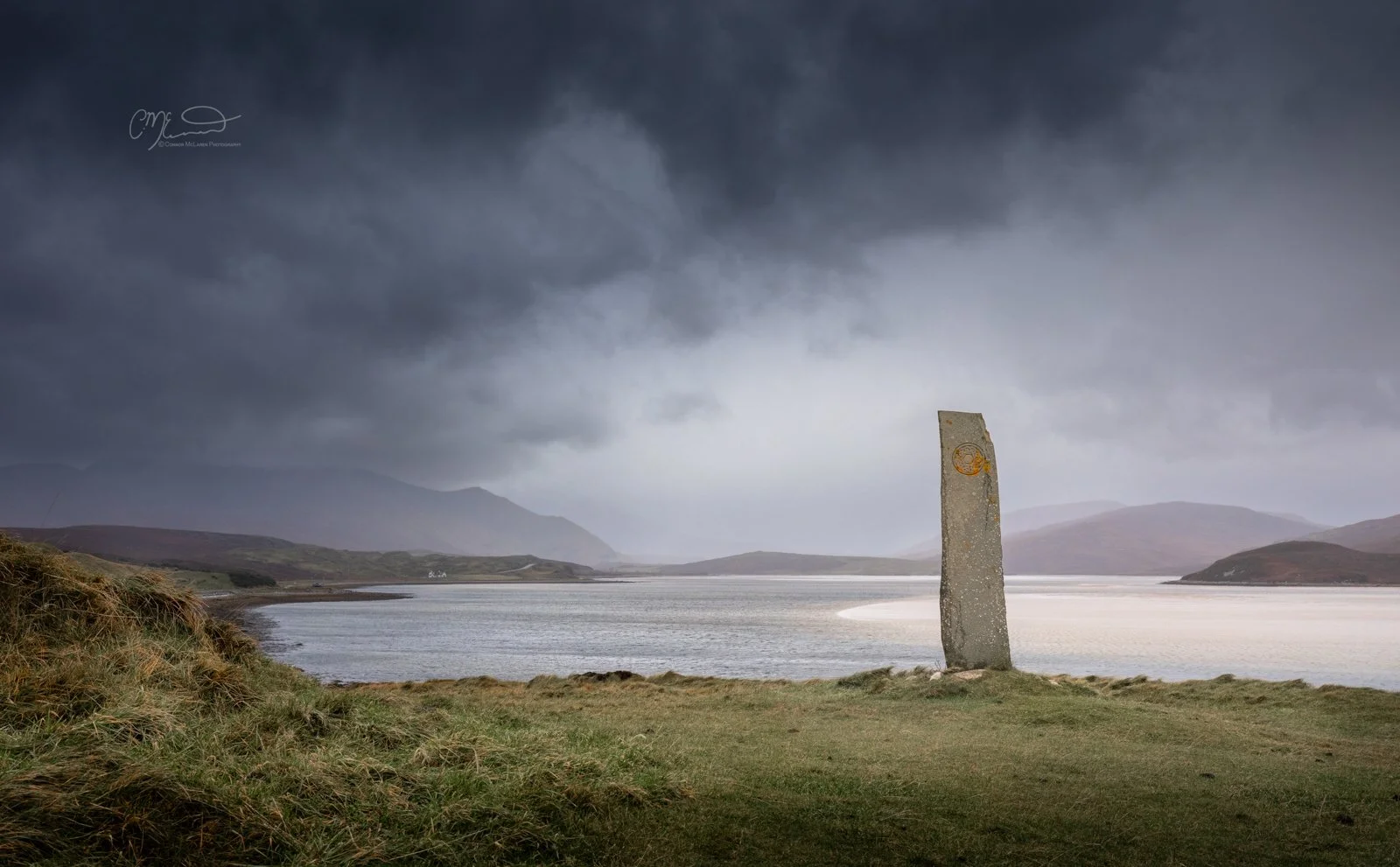 Scotland, north coast 500, NC500, durness. A tall, weathered stone marker standing by a lake surrounded by grassy land, mountains in the distance, and dark, moody clouds overhead.
